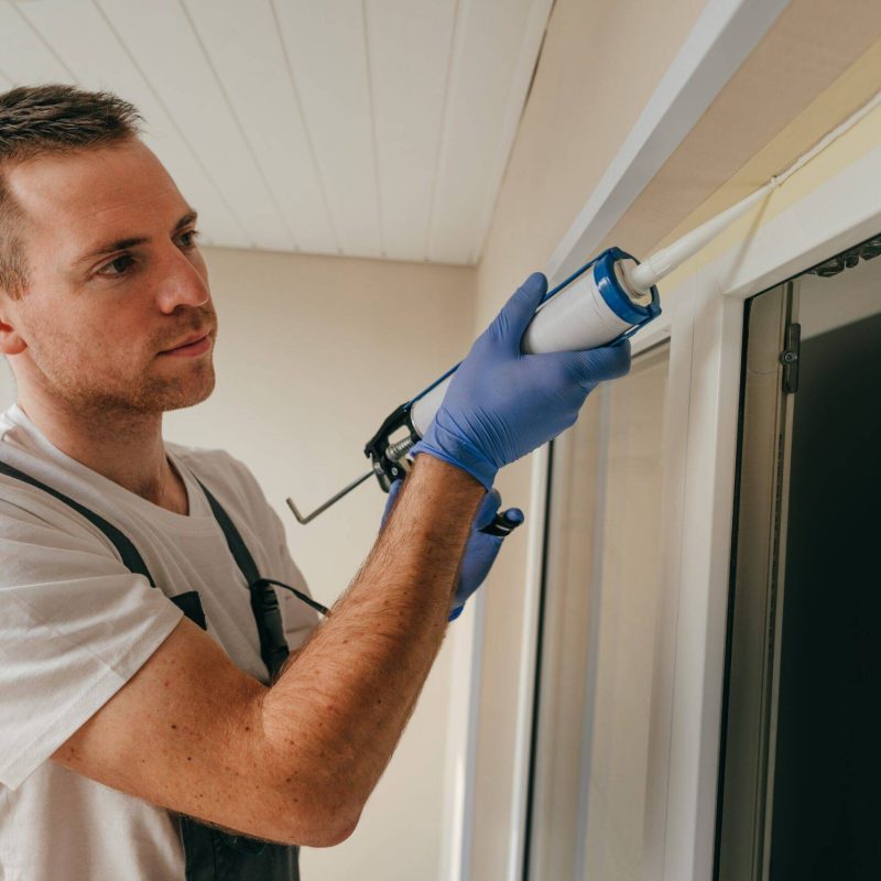 Young man wearing overalls sealing cracks between window and trim using waterproof silicone caulk on the balcony.